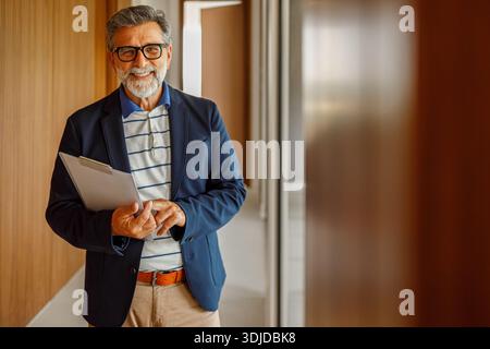 Ritratto di un uomo d'affari sorridente con barba che tiene in mano gli appunti e si trova davanti a un muro di legno in un ufficio moderno Foto Stock