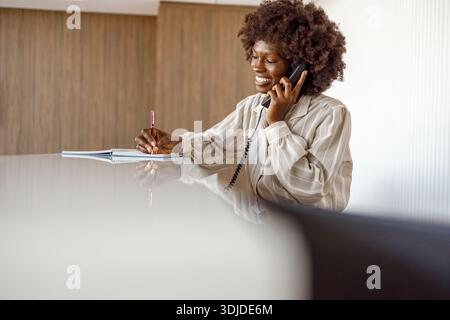 Una receptionist donna scrive appunti mentre parla al telefono e assiste gli ospiti al banco della reception dell'hotel Foto Stock