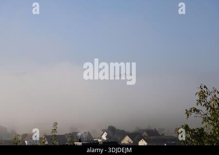 Mattinata di Foggy sopra un piccolo villaggio rurale con case che emergono dalla fitta nebbia. Paesaggio tranquillo e suggestivo con luce soffusa e atmosfera autunnale. Foto Stock