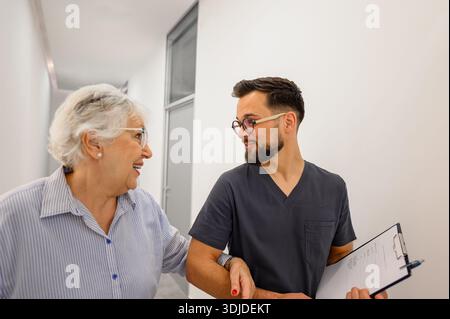 Paziente anziano e medico maschile che cammina braccio nel braccio e discute rapporti di test medici nella hall dell'ospedale Foto Stock