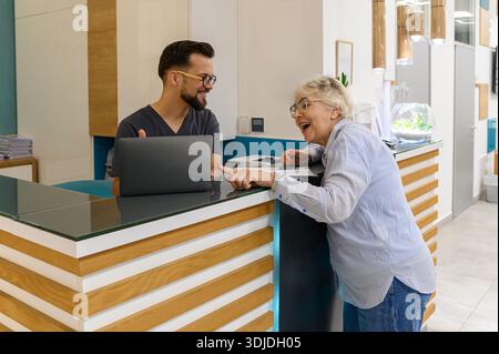 Donna anziana che guarda il computer portatile come il medico spiega i dettagli medici al banco di ricevimento in clinica oftalmologia Foto Stock
