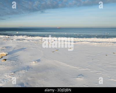 Ampia vista aerea di una spiaggia innevata del Mar Baltico in inverno con una nave da carico lontana sull'orizzonte blu freddo. Foto Stock