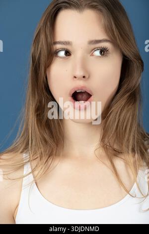 Giovane donna che mostra sorpresa e shock con la bocca aperta e gli occhi ampi su sfondo blu Foto Stock