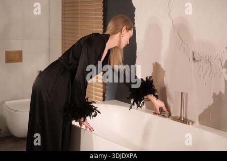 La giovane donna con l'abito nero regola il rubinetto per riempire un'elegante vasca da bagno in un bagno contemporaneo con persiane in legno e tonalità neutre con copia Foto Stock