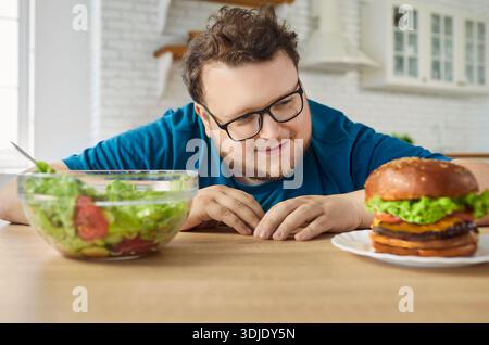 Uomo affamato che sceglie l'hamburger anziché l'insalata, scelta dietetica di persona in sovrappeso divertente Foto Stock