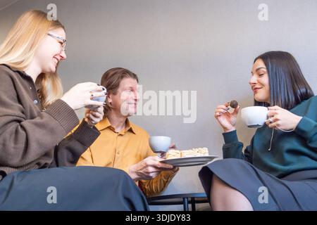 Un uomo e due giovani donne condividono un momento di luce con caffè e pasticcini in un ambiente interno rilassato, che mette in mostra un'atmosfera amichevole con spazio per fotocopie Foto Stock