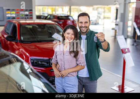 Bella coppia felice e amorevole che acquista una nuova auto in salone, in piedi sopra un'auto di lusso, abbraccia e mostra le chiavi della loro nuova auto, copia spazio Foto Stock