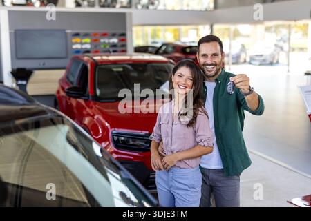 Bella coppia felice e amorevole che acquista una nuova auto in salone, in piedi sopra un'auto di lusso, abbraccia e mostra le chiavi della loro nuova auto, copia spazio Foto Stock