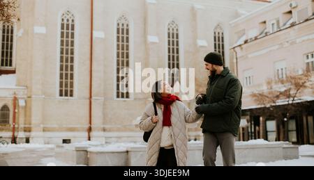 Una donna coreana con una sciarpa rossa ride e si tiene per mano un uomo caucasico. Sono in una piazza innevata con un grande edificio dietro di loro. Sembra li Foto Stock