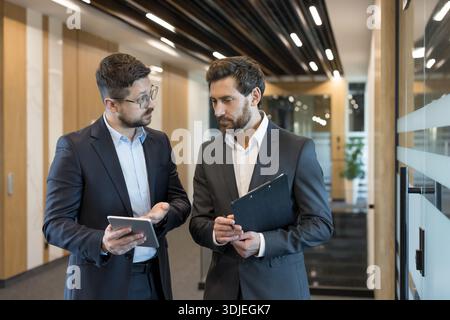Due uomini d'affari attraversano un moderno corridoio d'ufficio, condividendo idee da un tablet e da appunti mentre discutono strategia e lavoro di squadra in un ambiente aziendale professionale Foto Stock