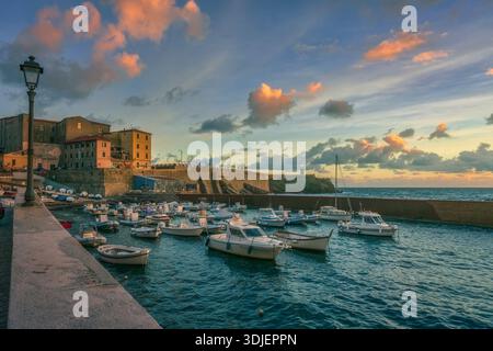 Vista delle piccole imbarcazioni ormeggiate nel porto di Piombino. Piazza Bovio storica e il suo faro sullo sfondo sotto un colorato cielo al tramonto, in Toscana, Foto Stock