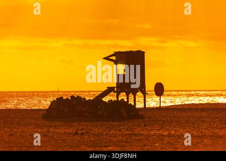 La torre del bagnino è caratterizzata da un vivace tramonto arancione su una spiaggia sabbiosa. Un mucchio di rocce e un anello rosso si trovano in primo piano vicino all'oceano i. Foto Stock