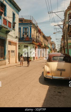 L'Avana, Cuba 2024 Apr16 - i residenti locali camminano attraverso un tipico quartiere fiancheggiato da colorati edifici coloniali e da un'auto d'epoca lungo la strada Foto Stock