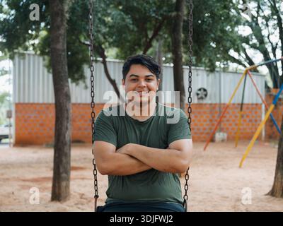 Ritratto di un giovane uomo seduto su un'altalena, braccia incrociate e sorridente alla telecamera in un parco con alberi e parco giochi sullo sfondo. Immagine ideale per Foto Stock
