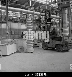 Sloviansk, Ucraina, URSS - anni '1980: Una donna che guida carrelli elevatori trasporta un massiccio isolante in ceramica ad alta tensione all'interno dell'officina dell'impianto isolante ad alta tensione di Sloviansk (ZVI). Un altro lavoratore sta su una scala che regola la colonna in porcellana vicino ai macchinari industriali. Questa autentica fotografia d'archivio in bianco e nero cattura la scala dell'industria pesante sovietica e la meccanizzazione della logistica. L'immagine documenta il lavoro quotidiano dei dipendenti delle fabbriche nella pacifica regione del Donbas molto prima della guerra. Foto Stock