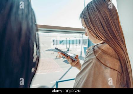 Due giovani donne stanno aspettando in aeroporto. Stanno guardando fuori dalla grande finestra. Uno ha lunghi capelli castani, l'altro ha lunghi capelli neri. Foto Stock