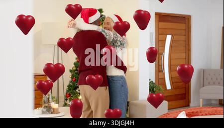 Abbraccia una coppia che indossa cappelli di Babbo Natale e maglioni accoglienti nel soggiorno, con decorazioni a cuore rosso Foto Stock