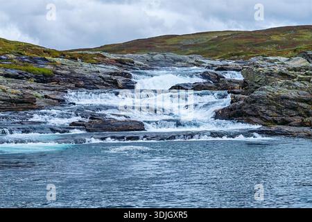 Il paesaggio di Vildmarksvagen raffigura un potente fiume che forma piccole cascate su numerose rocce scure, con vaste colline coperte da vegetazione sparsa Foto Stock