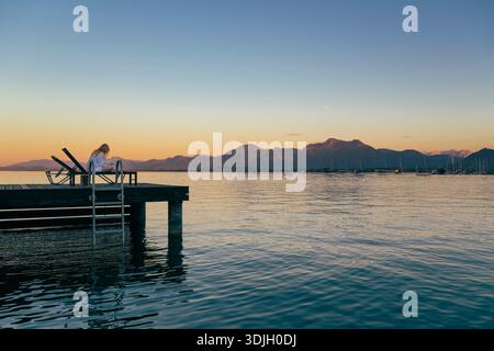 Una giovane donna in accappatoio bianco giace su una sdraio su un molo del lago Chiemsee al crepuscolo, leggendo un libro, Baviera, Germania Foto Stock