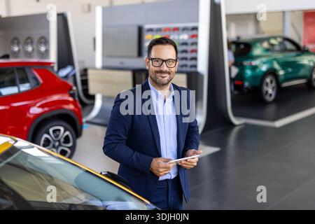 Bell'aspetto, allegro e cordiale venditore posa in un salone di auto o in uno showroom e guarda la fotocamera. Foto Stock