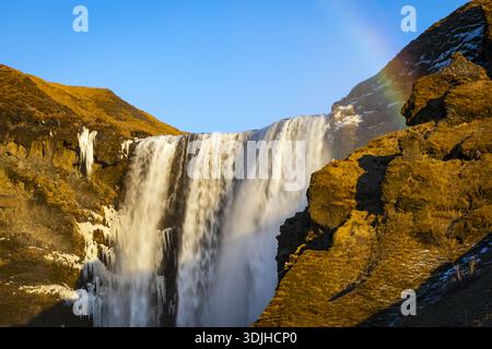 Vista della maestosa cascata di Skógafoss che si tuffa dalle aspre scogliere sotto un cielo limpido, un vivace arco arcobaleno che aggiunge magia, Skogafoss Wate Foto Stock