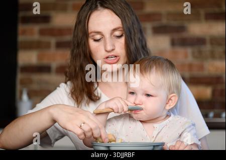 Allattamento del bambino. La madre insegna al bambino tecniche di alimentazione adeguate. La donna aiuta il bambino ad assaggiare il cibo, con cucchiaio e piatto, e guida il bambino durante l'uso dei pasti Foto Stock