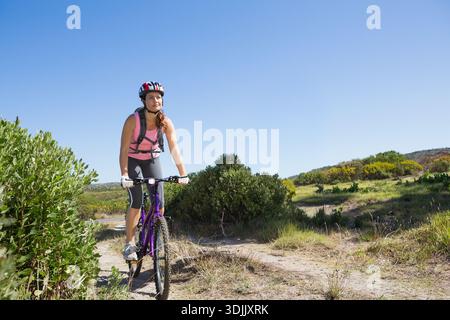 Donna che guida una mountain bike viola su una stretta pista sterrata con casco rosso bianco guanti e zaino. Ciclista, all'aperto, avventura, percorso, semiarido, nat Foto Stock