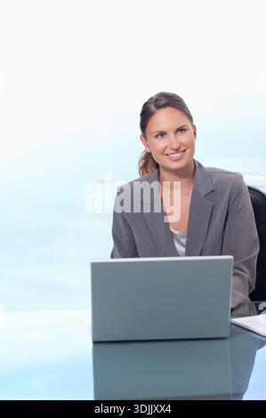 Donna che indossa un blazer grigio seduto a una scrivania di vetro in un ufficio moderno a scrivere su un notebook vicino al notebook. Minimalista, con finestre, luce naturale, lungomare, sereno, f Foto Stock