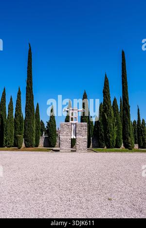 Moderna scultura in pietra all'ingresso di un cimitero, fiancheggiata da alti cipressi. La città vecchia di Labin è visibile in lontananza sotto un cielo blu. Foto Stock
