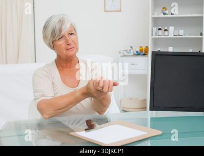 Donna anziana che tiene il polso durante la consulenza in sala esame medica con appunti su scrivania di vetro. Clinica, paziente, appuntamento, benessere, calma, minimali Foto Stock
