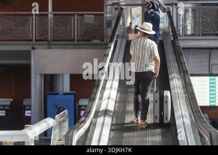 Donna con un cappello che tira i bagagli su una scala mobile, che viaggia attraverso un moderno terminal aeroportuale per le vacanze, Spagna, Madrid Foto Stock