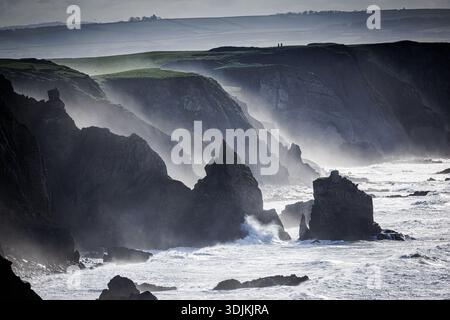 Due camminatori sulla cima di una scogliera torreggiante mentre i mari selvaggi si schiantano contro le rocce sul sentiero della costa sud-occidentale a North Conrwall, vicino a Bude e Sandymouth Foto Stock