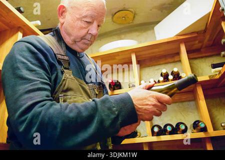 Un uomo anziano esamina attentamente una bottiglia di vino in una cantina rustica, con scaffali in legno che espongono una serie di bottiglie di vino, sottolineando la passione per Foto Stock
