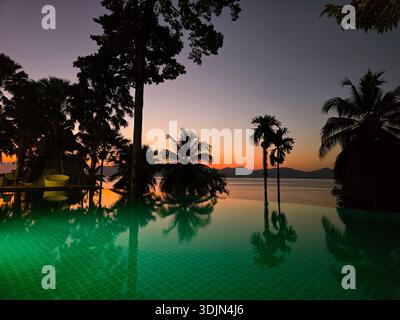 Lo spettacolare tramonto dipinge il cielo sull'isola di Koh Phayam, con palme che ondeggiano dolcemente accanto alla piscina a sfioro. Le acque tranquille riflettono tonalità vibranti, cr Foto Stock