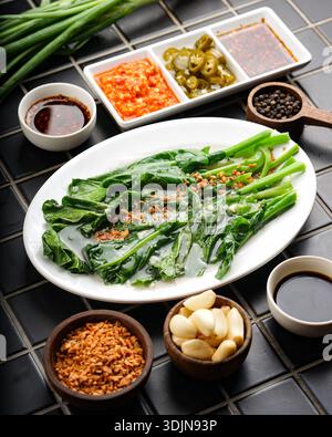 Verdure fresche e verdeggianti servite con varie spezie ed erbe aromatiche su un tavolo con piastrelle nere, creando una vivace e appetitosa cucina di ispirazione asiatica. Foto Stock