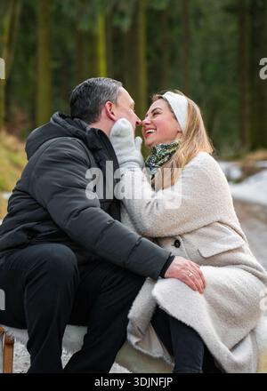Uomo e donna felici e romantici seduti su una slitta, si abbracciano dolcemente e si guardano negli occhi sul sentiero innevato della foresta. Coppia innamorata che si gode una vacanza invernale Foto Stock