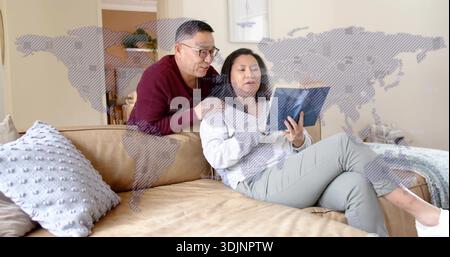 Uomo anziano pendente che indossa un maglione e una donna che tiene un tablet sul divano di casa, sovrapposizione di mappe Foto Stock