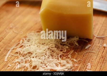 Primo piano del pelo di formaggio duro appena grattugiato e del blocco di formaggio su un tagliere di legno. Consistenza degli ingredienti. Foto Stock