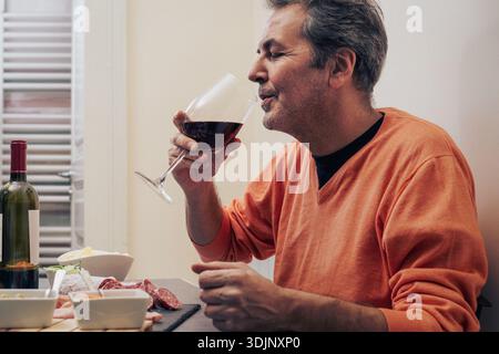 Un uomo maturo con un maglione arancione pratica una sapiente degustazione di vino rosso mentre si gusta un momento slow food con antipasti a casa, illustrando l'intento Foto Stock