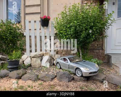 Un'auto giocattolo Mercedes-Benz SLR McLaren color argento è collocata in un giardino tra rocce e cespugli verdi accanto a una recinzione di legno bianca e all'ingresso di una casa Foto Stock