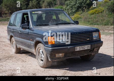 Vista frontale di un classico SUV di lusso Land Rover Range Rover P38A blu polveroso (seconda generazione, modello 1995-2002) che guida su una strada sterrata sotto un luminoso su Foto Stock