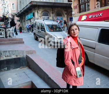 Donna adulta che viaggia da sola, esplora una strada della città di Istanbul, indossa una giacca e porta uno zaino, sorride alla macchina fotografica, con edifici urbani A. Foto Stock