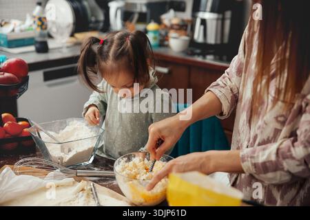 Una madre e sua figlia lavorano insieme in una cucina. La figlia mescola gli ingredienti in una ciotola mentre sua madre la guida. Vari utensili da cucina e. Foto Stock