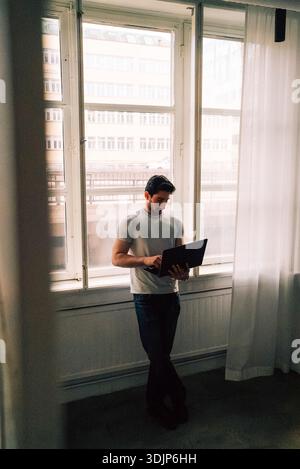 Vista ad angolo alto di un giovane professionista della tecnologia maschile che utilizza un computer portatile mentre si trova vicino alla finestra in ufficio Foto Stock