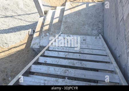 Vista dall'alto verso il basso della ripida scala in legno che conduce alla piattaforma in calcestruzzo nel cantiere, con un'ombra luminosa che getta luce solare sullo spettacolo della struttura non finita Foto Stock