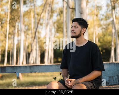 Ritratto di un giovane uomo seduto su una panchina in un parco alberato, con postura rilassata e che guarda di lato. Immagine adatta ai concetti di tranquillità, Foto Stock