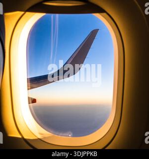 Ala dell'aeroplano vista dal finestrino del passeggero durante il volo al tramonto, con bordo dorato che si illumina contro cieli blu e nuvole lontane Foto Stock