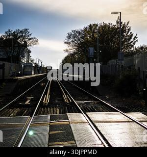 Binari ferroviari che conducono attraverso un passaggio a livello in una tranquilla stazione suburbana in Inghilterra al crepuscolo, con alberi sagomati e piattaforme che creano un forte Foto Stock