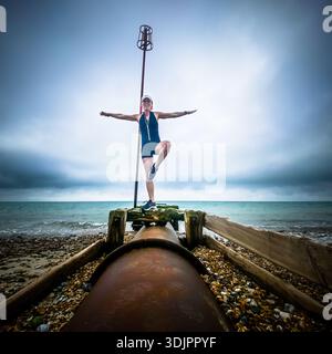 Donna che pratica yoga in posa sugli alberi su una pipa arrugginita tra ciottoli su una spiaggia inglese, contro un orizzonte marino nuvoloso Foto Stock