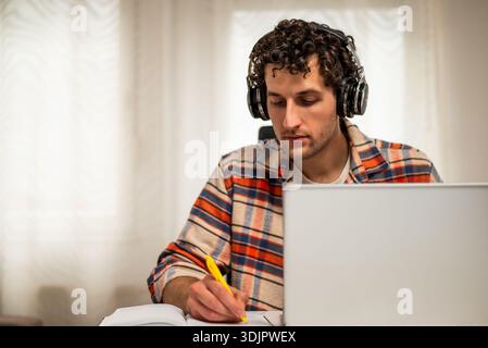Giovane uomo concentrato che indossa le cuffie wireless, lavora sul suo notebook a casa mentre prende appunti su un notebook. Foto Stock